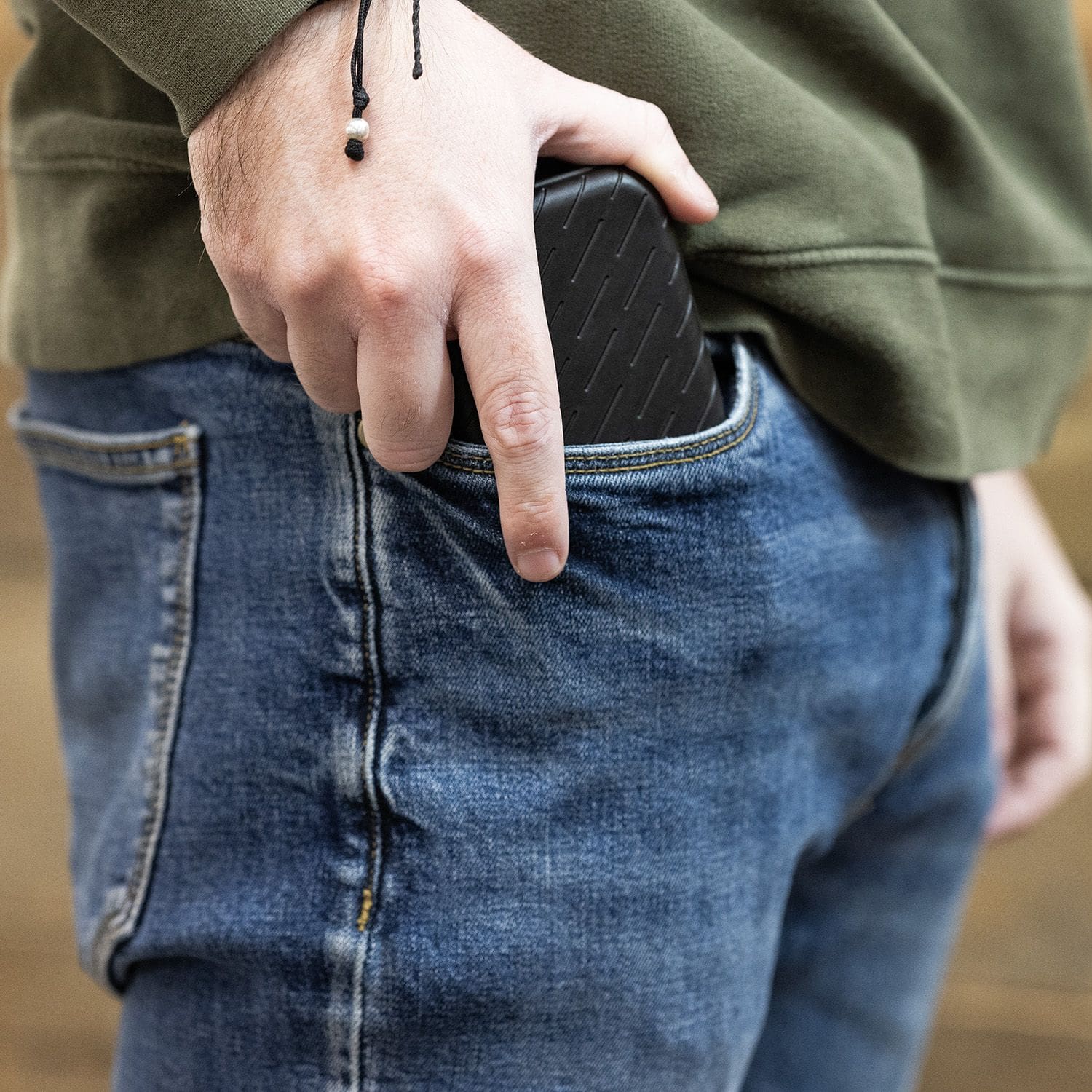 Harrows Slimcase Dart Case Eine Person steckt ein schwarzes Etui in die Hosentasche. Sie trägt eine grüne Jacke und eine blaue Jeans.
