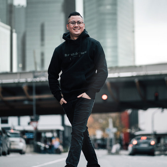 Barrels and Shafts Hoodie - Black Eine Person trägt einen schwarzen "Barrels and Shafts" Hoodie und steht entspannt auf einer Straße. Im Hintergrund sind unscharfe Gebäude und Autos zu sehen.