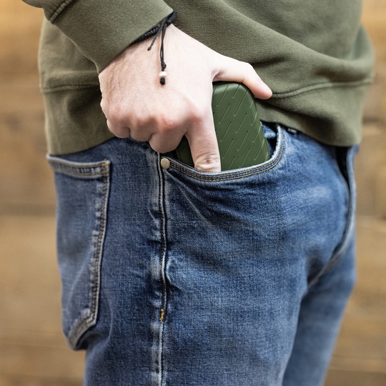 Eine Person steckt ein grünes Etui in die vordere Hosentasche einer Jeans. Das Etui hat eine strukturierte Oberfläche und passt gut in die Tasche.