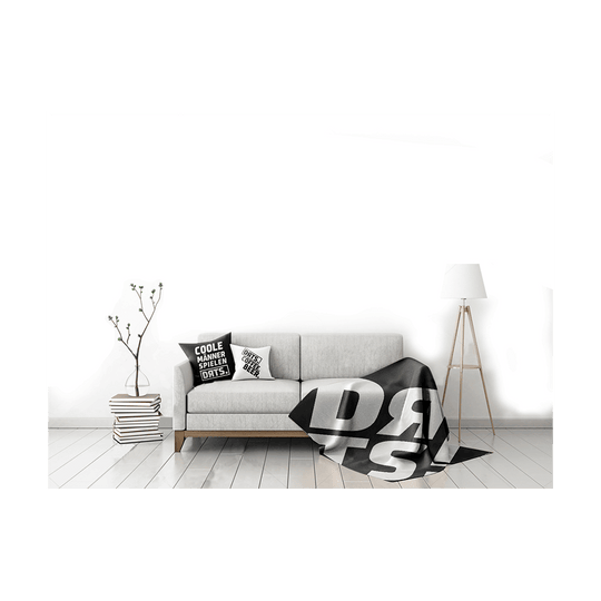 DRTS_K1_DRTS_Kissen_Schwarz_3 Das Bild zeigt ein modern eingerichtetes Wohnzimmer mit einem hellgrauen Sofa. Auf dem Sofa liegt das Produkt "SPORT1 DRTS. Kissen" mit auffälligem schwarzen und weißen Schriftzug.